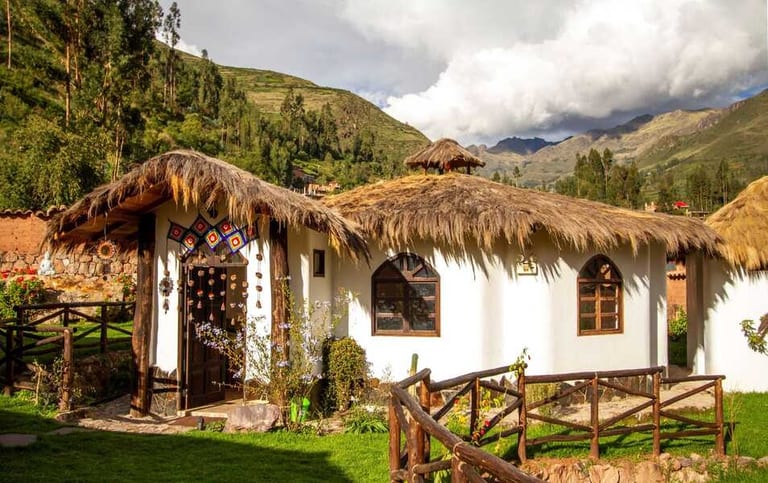 A white-walled bungalow with a thatched roof at Ayni Sanctuary, Calca, surrounded by green lawns and Andean mountains under a cloudy sky.